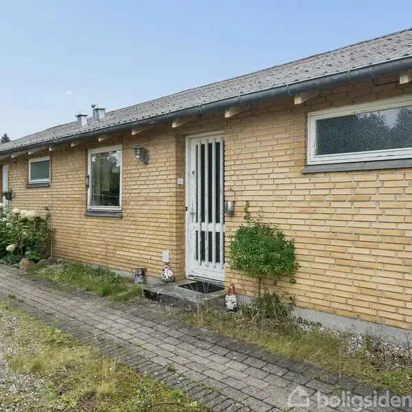 En langstrakt, gulstensbolig med hvid dør og vinduer. Omgivet af en flisebelagt sti og lav beplantning. Skyfri himmel anes over bygningen.