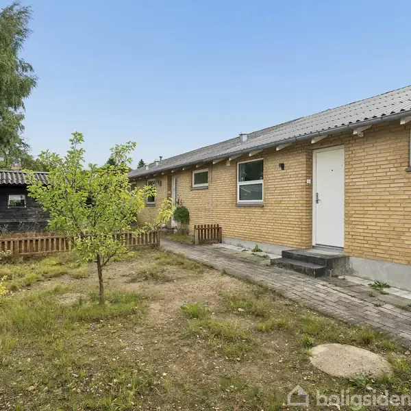 Et enkelt hus af gule mursten med en hoveddør og små vinduer; en beskeden have foran med en græsplæne og træer, og et lille skur omkranset af et hegn.