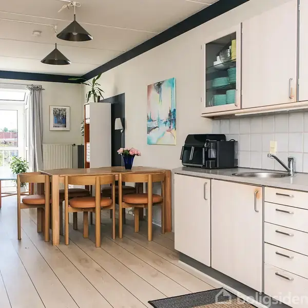 Køkken-alrum med hvidt køkkenskab til højre, ovn og kaffemaskine. I midten står et spisebord med brune stole. Bagerst er der en sofa ved en stor glasdør med udsigt til haven.
