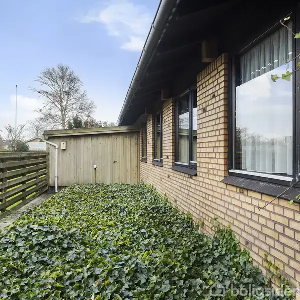 Murstenshus med store vinduer er omgivet af en frodig have med efeu. En træhegn løber langs siden, og en lille træskur ses foran. Himlen er blå med skyer.