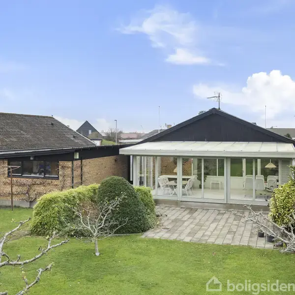 Et hus med glasindhegnet terrasse står stille. Haven omkring er velplejet med græs og buske. Klar blå himmel i baggrunden.