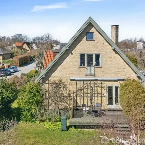Et A-formet hus med en terrasse foran, omkranset af en have med buske og træer. Det ligger i et villakvarter med andre huse og en vej i baggrunden.