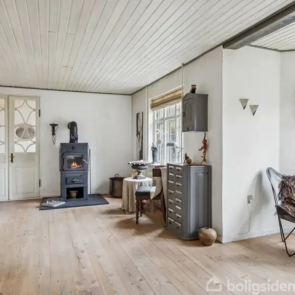 Et lyst rum med trægulv indeholder en brændeovn, der står langs væggen, ved siden af en vitrine, et lille bord og en stol med et tæppe.