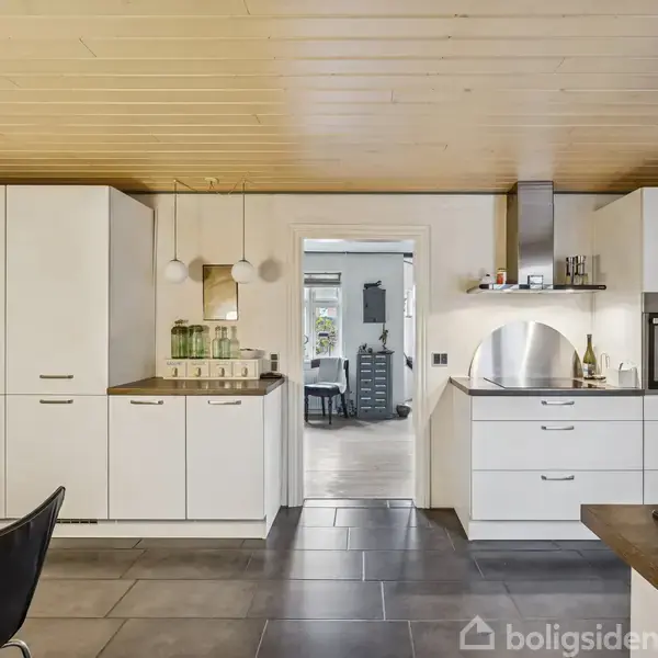 Et moderne køkken med hvide skabe og skuffer, en indbygget ovn og emhætte. I midten hænger tre lamper over en bordplade. Baggrunden viser en åben dør til næste rum.