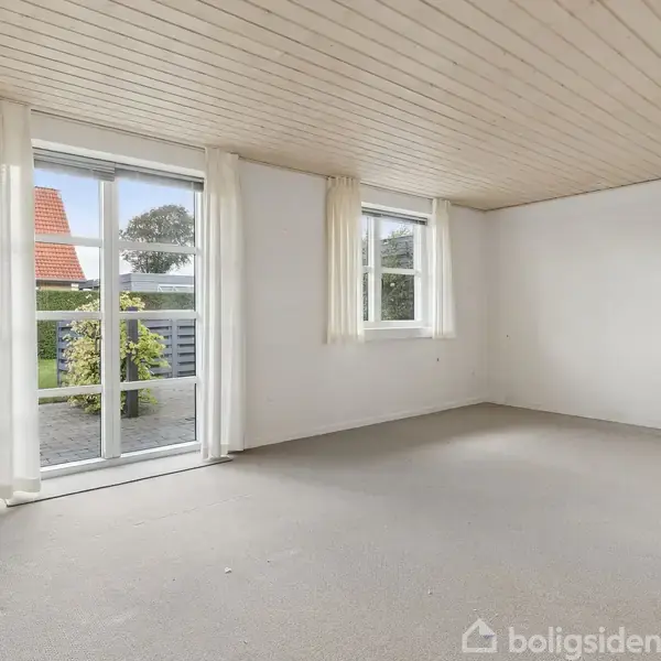 Et lyst, tomt rum med lyse vægge og loft. Store hvide gardiner hænger ved glasdøre og et vindue, der åbner ud mod en have med et rødt tag i baggrunden.