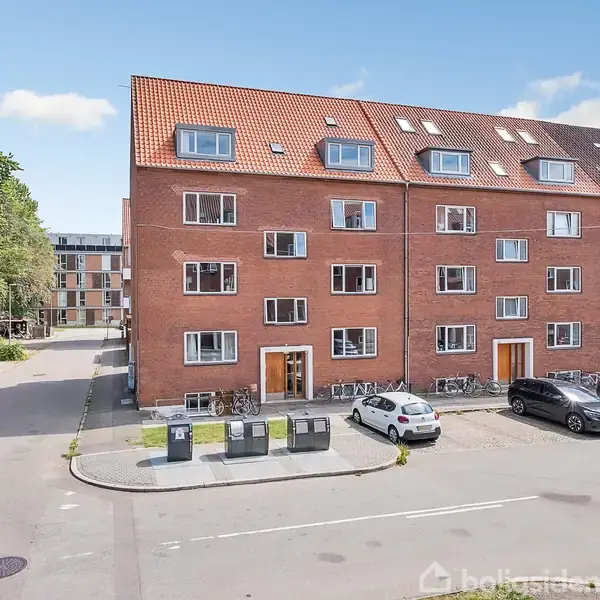 Rød murstensbygning står stille langs vejen med parkerede biler foran. Til venstre er træer og en sidevej, og i baggrunden ses lejlighedskomplekser.