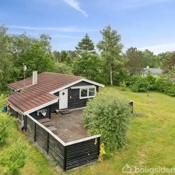 Et sort træhus med hvide vinduer og skorsten står i en frodig have omgivet af høje træer og grønne buske. Græsplænen omgiver huset uden andre bygninger i nærheden.
