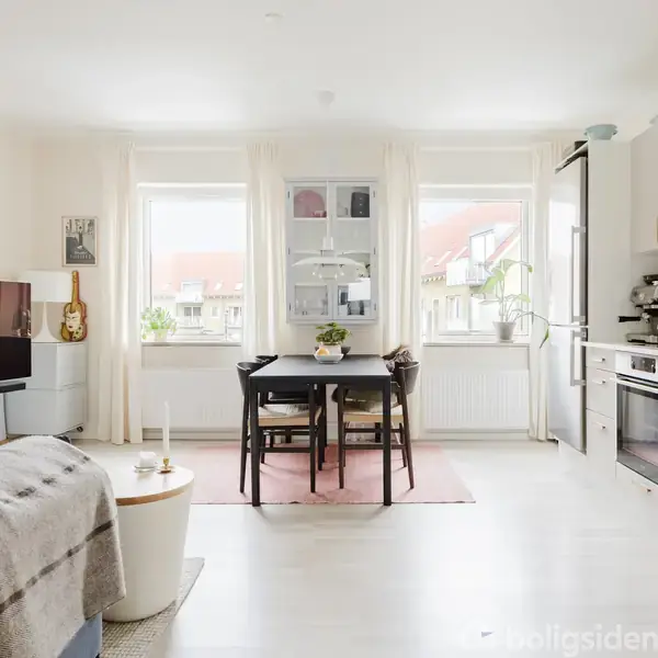 Stuen har en fladskærms-TV mod en væg, borde står i midten, omgivet af stole, med lysindfald fra vinduerne. Højre side har et køkken med moderne apparater.