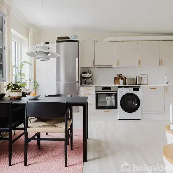 Et lyst køkken-alrum med spisebord og fire sorte stole på et lyserødt tæppe. Køleskab, vaskemaskine, ovn og skabe er placeret i baggrunden. En lille plante står på bordet.