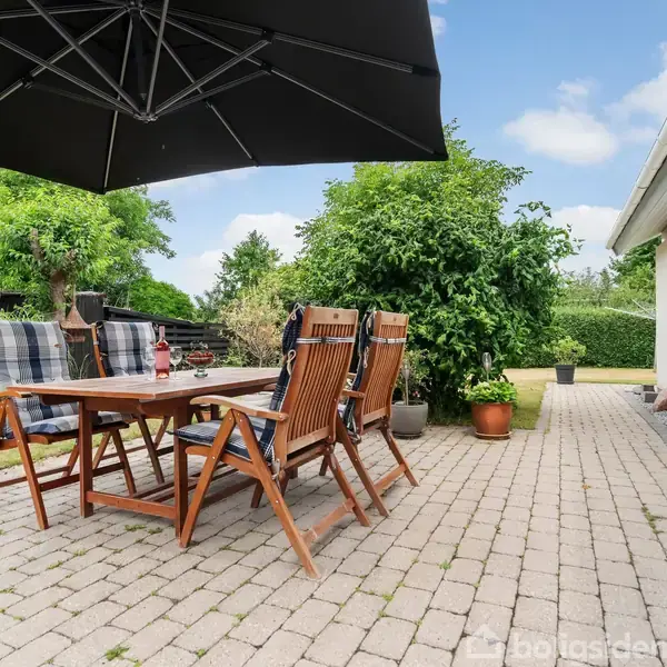 Terrassemøbler står under en stor, sort parasol på en flisebelagt terrasse ved siden af et hvidt hus. Omkring er der planter, buske og en lille havefigur i baggrunden.
