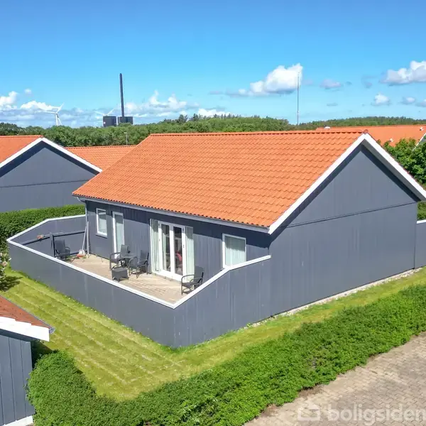 Gråt hus med rødt tag står stille omkranset af hæk og græsplæne. Huset har en terrasse med stole, og flere lignende huse er synlige i baggrunden under en blå himmel.