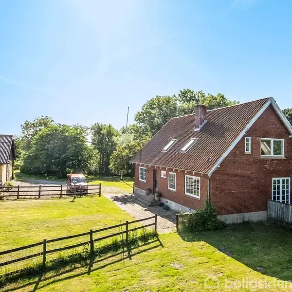 En rød murstensbygning står stille på en solrig dag, omgivet af en grøn græsplæne og træer. Ved siden af ses et hegn og en gul bygning.