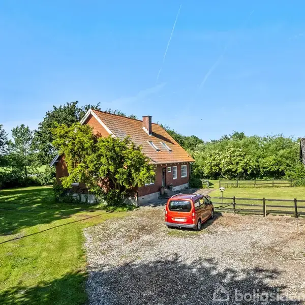 Et rødt murstenshus står stille i en grøn have, omgivet af træer. En rød bil er parkeret på en grussti ved siden af en lade under en solrig blå himmel.