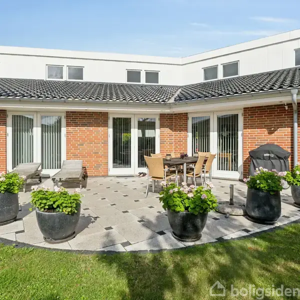 Rund terrasse med fliser, omkranset af sortmalede krukker med grønne planter. Havemøbler står midt på terrassen, foran et murstenshus med store vinduer og sorte tagsten.