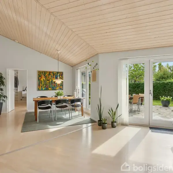 Et lyst og rummeligt opholdsstue, hvor et spisebord med stole står på et gulvtæppe. Store vinduer til højre åbner op til en terrasse og have med grøn bevoksning. Væggene prydes af farverige kunstværker.