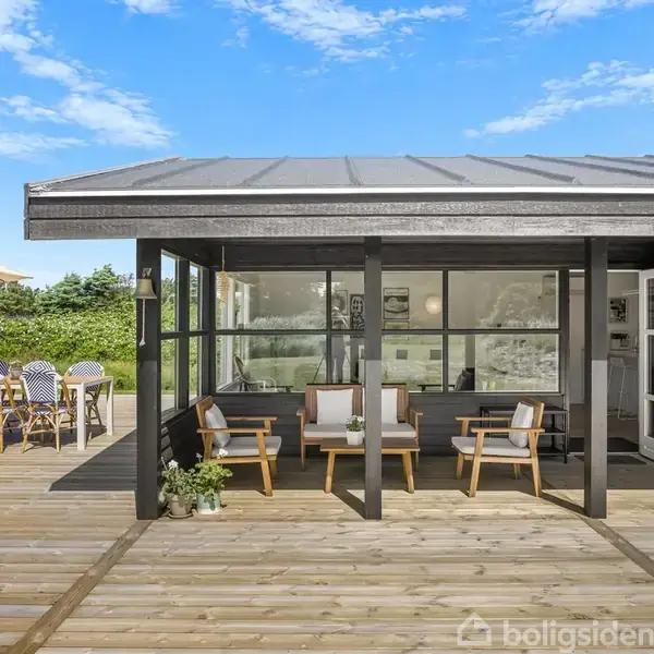 Terrasse med træmøbler og planter står på en stor træplatform ved et sort hus med store vinduer. I baggrunden er en parasol over et spisebord, omgivet af grønne buske.