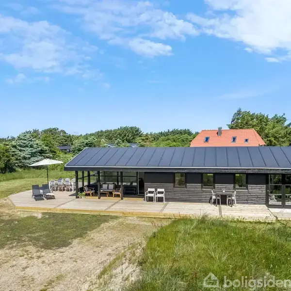 Et sort træhus står stille på en træterrasse. På terrassen er der havemøbler og en parasol. Omgivelserne er grønne, med buske og et andet hus med rødt tag i baggrunden.