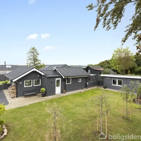 Gråmalet hus med sorte tage står midt i en velholdt have. Der er enkelte unge træer plantet i forgrunden, og i baggrunden ses skov og andre bygninger.