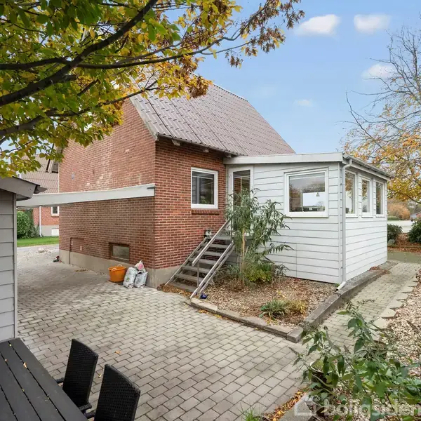 Rødt murstenshus med baghave og vinterhave, omgivet af fliser og havebede. En terrasse med borde og stole ses til venstre. Løvet på træerne er begyndt at falde.