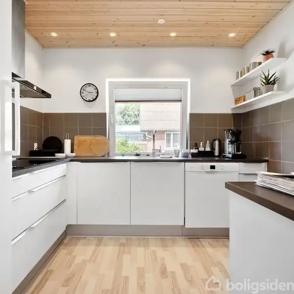 Et moderne køkken viser hvide skabe og apparater. Rummet er lyst med trægulv og loft. Udenfor ses et vindue med en haveudsigt. En vægur hænger på væggen.