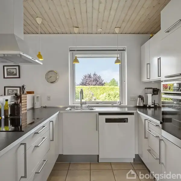 Køkken med hvide skabe og sort bordplade udstyret med ovn og kogeplade. Vægur og malerier hænger på væggen. Vindue med udsigt til en grøn have.
