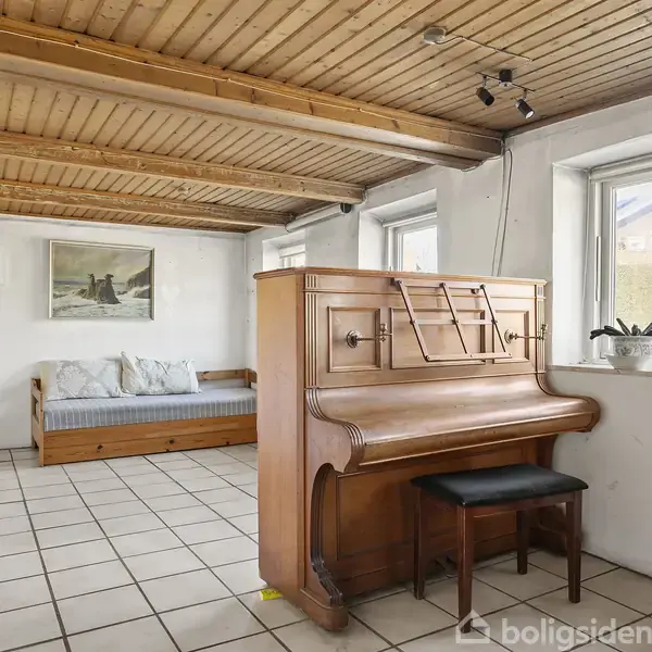 Et klaver står i et rum med hvidmalede vægge og flisegulv, ved siden af en bænk ved vinduet og en daybed i baggrunden.