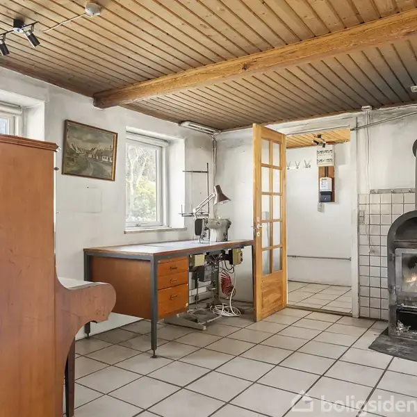 Brændeovn står i hjørne og opvarmer rum med hvide vægge og træloft. Et skrivebord med en lampe ved siden af åbner til et tilstødende rum gennem en glassdør.