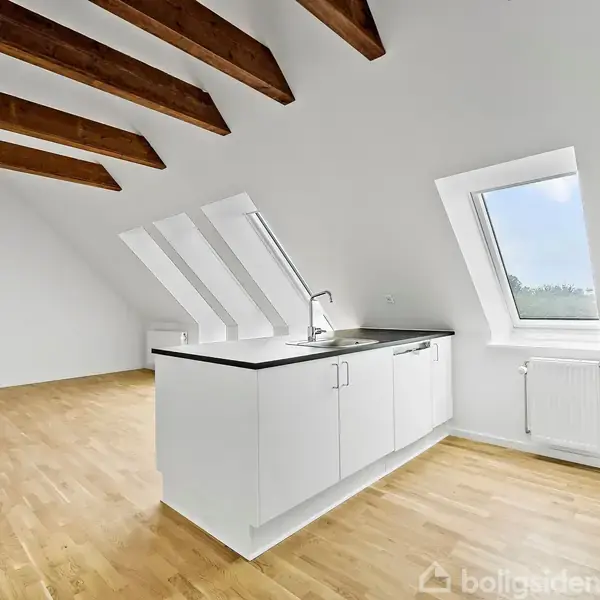 Køkken med hvidt skab og sort bordplade står under skrånende vinduer. Loftsbjælker i træ pyntet over lyse trægulve. Rummet er lyst og rummeligt med hvidmalede vægge.