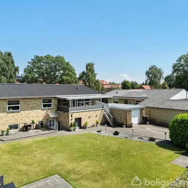 En murstensbygning med fladt tag står omgivet af have og terrasse; en grusbelagt indkørsel leder til en garage. Omkring huset ses grønne buske og træer under en blå himmel.