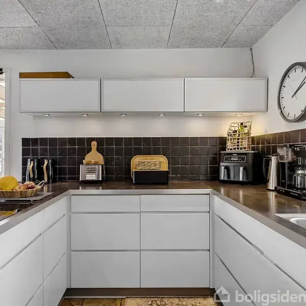 En moderne køkkenvask i et lyst køkken. Skabene er hvide, bordpladen grå. På væggen hænger et ur. Små apparater og frugter er placeret på bordet.