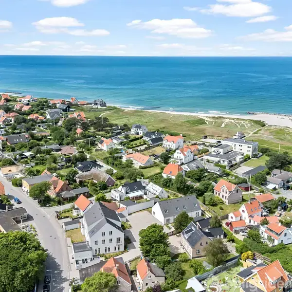 Kystby med røde tage ligger langs en vej ved havet. Huse er spredt ud i et grønt område, mens det blå hav strækker sig til horisonten under en klar himmel.