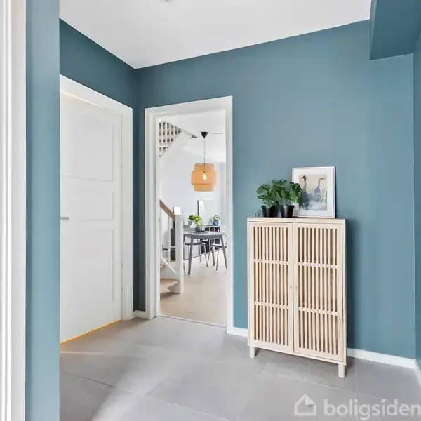 Et skab står op ad en blå væg, dekoreret med en plante og et billede. Gennem en åben dør ses et lyst rum med en bordlampe, stole og vindue i baggrunden.