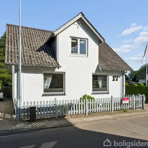 Hvidt hus med gråtag, to vinduer foran. Lille have og hvidt stakit. Husnummer 17. Solrig dag med få skyer.