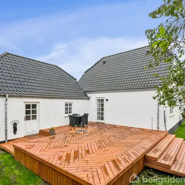 Huset har hvide vægge og gråt tegltag, der står på en stor træterrasse med borde og stole. Den grønne have omgiver huset, og himlen er blå.
