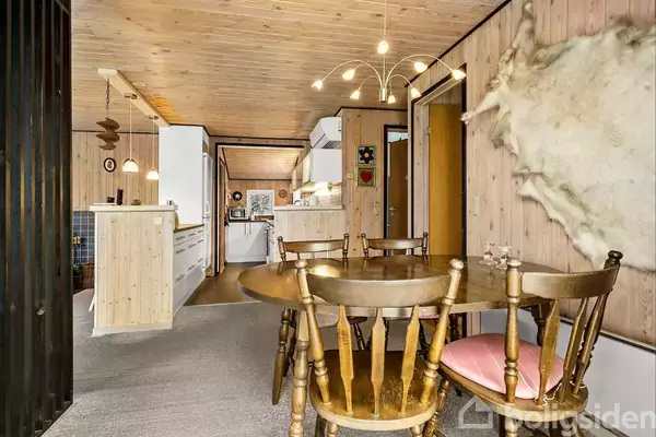 Rundt spisebord med fire træstole står i et træindrettet køkkenalrum. En moderne lysekrone hænger over bordet. På væggen hænger et hvidt dyreskind.