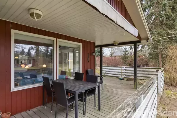 En sort spisebord med fire stole står på en træterrasse ved et rødmalet hus. Terrassen er omgivet af en lav træværn og der er buske i baggrunden.