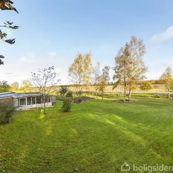En lille bygning står stille i en grøn have med nyslået græs, omkranset af træer og buske. En lys himmel oplyser landskabet.