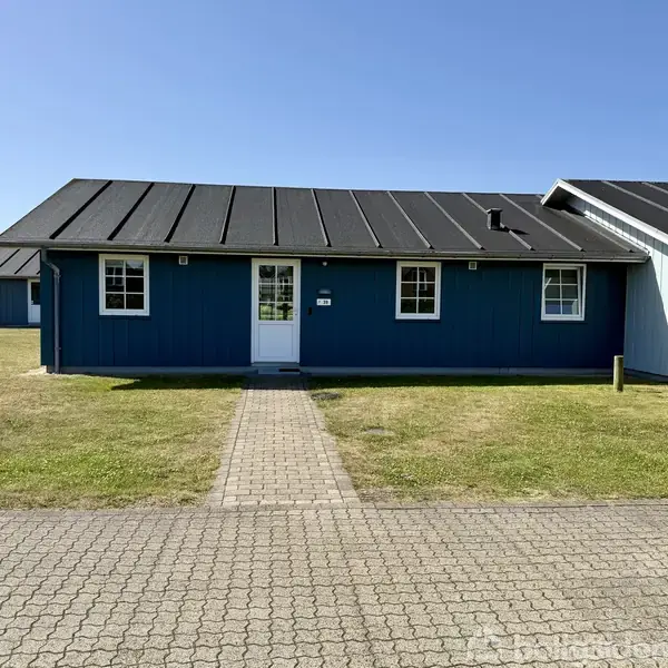 Blåt træhus står stille, omgivet af en plæne og en flisegang foran. Huset har et sort tag og hvide vinduesrammer. Det er solskin og himlen er klar.