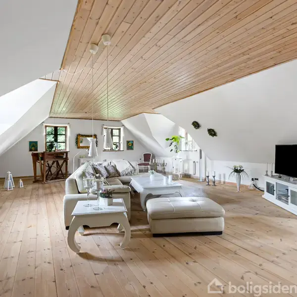 Hvid lædersofa står centralt på et lyst trægulv i en rummelig stue med skrålofter og et stort tv på en skænk. Grønne planter og moderne indretning skaber en hyggelig atmosfære.