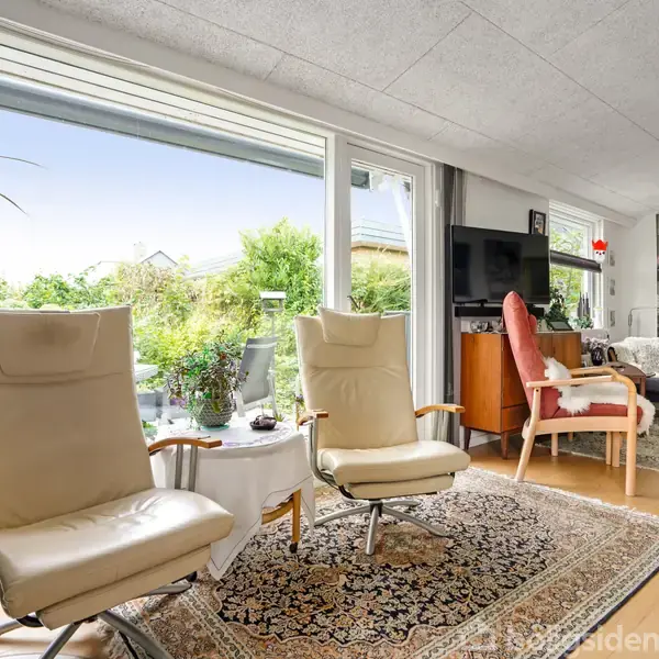 To lænestole står foran store vinduer med udsigt til en have. Indendørs er der et tæppe, stueplanter og sofaer i et lyst, indbydende rum.