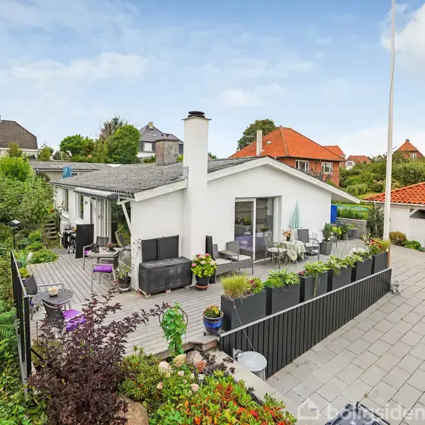 Køkken med hvidt skab og ø, indeholder køkkenudstyr og planter. To pendellamper hænger fra loftet. Store vinduer giver udsigt til en grøn have i baggrunden.