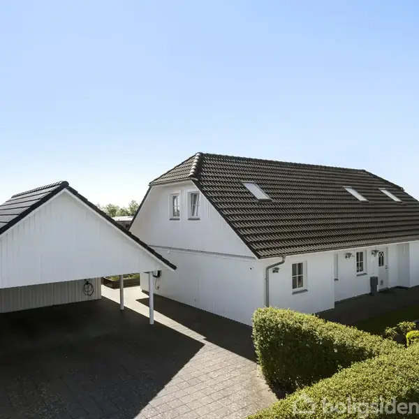 Hvidt hus med mørkt tag står i sollys. På højre side er en garage med åbent tag. Omkring er der klippede hække og en gårdsplads af sten.