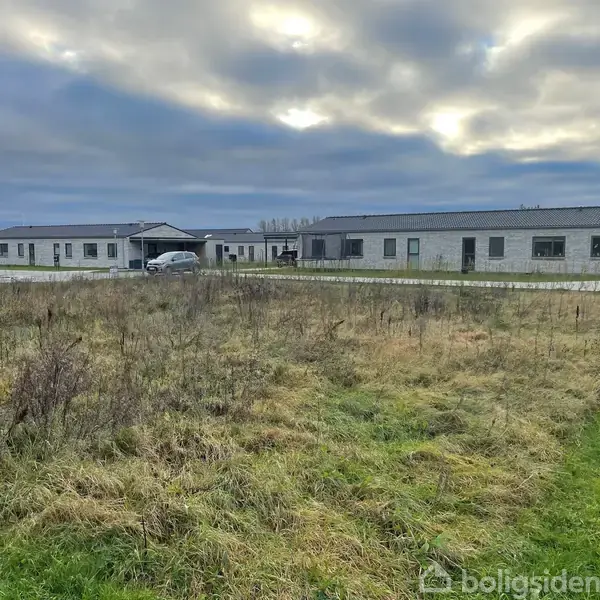 En åben græsmark med enkelte buske foran en række moderne enfamiliehuse i et boligområde. Biler er parkeret ved husene, og skyer dækker dele af himlen.
