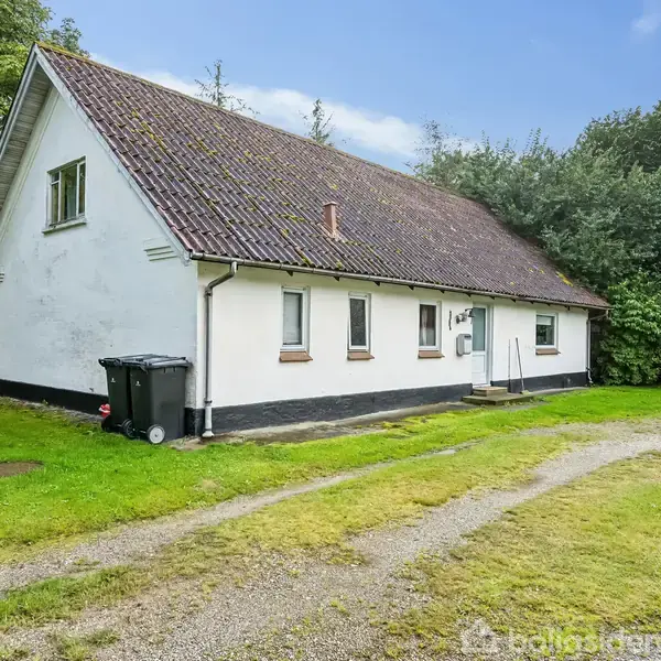 Et hvidt hus med brunligt tag står stille ved en grusvej. Omkring huset er der græs og træer. En sort skraldespand står ved siden af muren.