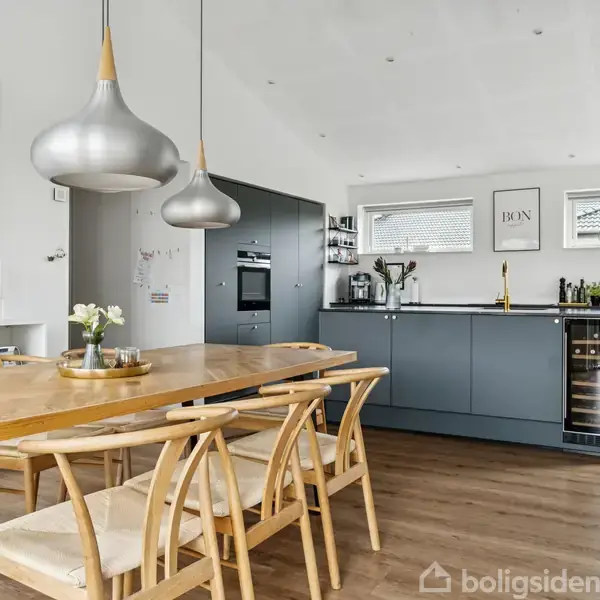 Træbord med seks stole står i et lyst køkken-alrum. Køkkenet har grå skabe, indbyggede ovne og en vinreol. Væggen er dekoreret med indrammede billeder og tekstplakater.