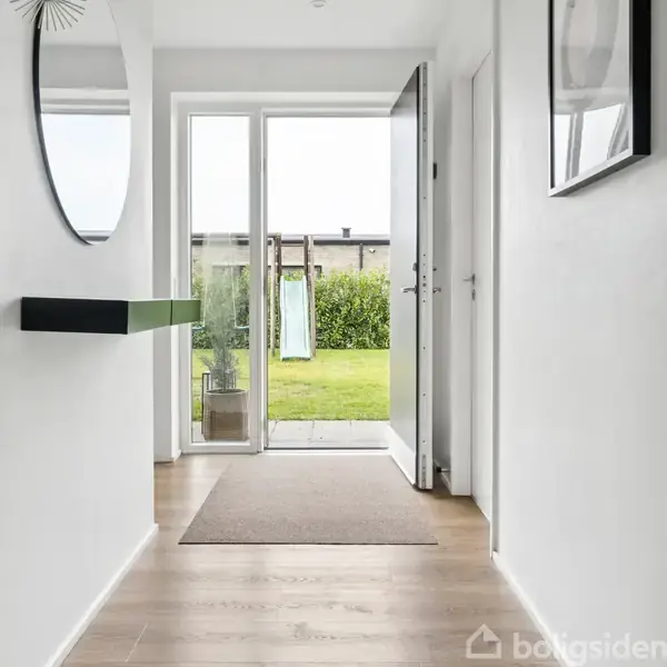 En gang med trægulv fører ud til en åben havedør, gennem hvilken en græsplæne og en lille rutsjebane kan ses. På væggen hænger en hylde og et spejl.