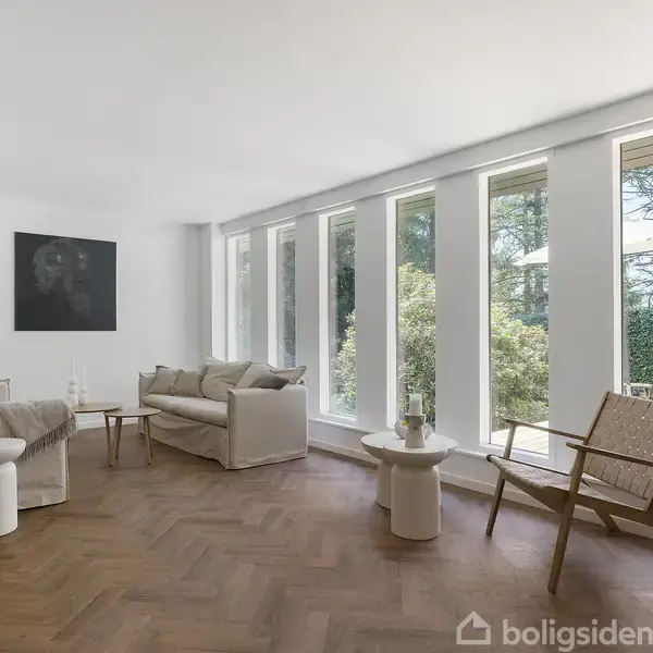 En lys stue med en sofa og lænestol placeret på et trægulv. Store vinduer åbner ud mod en have. Enkel indretning med et kunstværk og minimalistisk møblering.