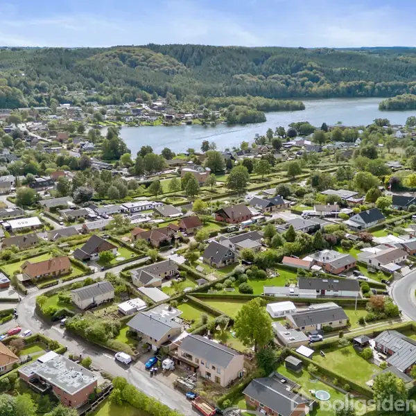 Boligområde med huse står stille i et frodigt landskab. En sø ses i baggrunden, omgivet af skovklædte bakker.