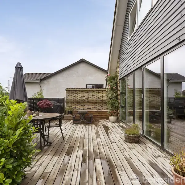 Træterrasse med havemøbler står foran et moderne hus med store vinduer. Planter og en parasol omgiver området, mens en murstensvæg ses i baggrunden.