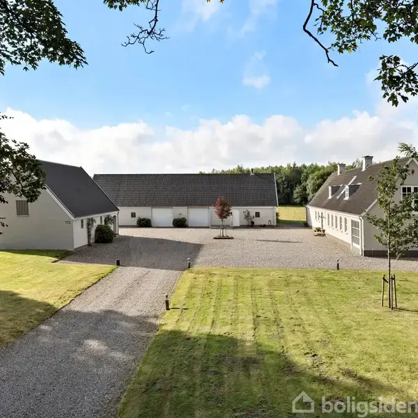En gårdsplads omgivet af tre hvide bygninger med sorte tage; grusbelægning forbinder dem. Græsplæne og træer omkranser området. Klar blå himmel i baggrunden.